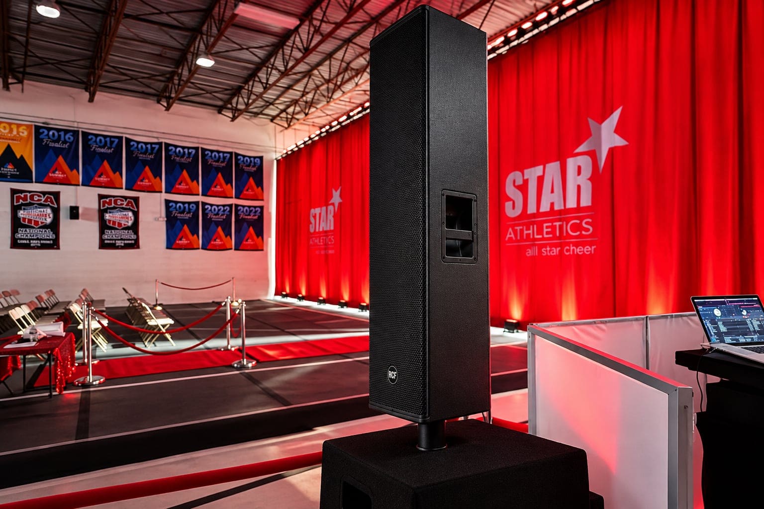 Speaker and subwoofer beside a cheer floor, spectator seating, and red draping